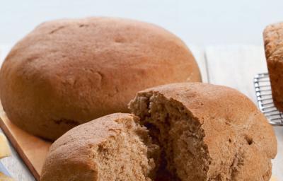 Fotografia em tons de marrom em uma bancada de madeira com uma tábua de madeira clara e o pão preto redondo grane apoiado nela, e um outro pedaço de pão já cortado. Fotografia em tons de marrom em uma bancada de madeira com uma tábua de madeira clara e o pão preto redondo grane apoiado nela, e um outro pedaço de pão já cortado.