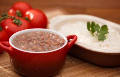 foto em tons de marrom, vermelho e branco de uma bancada marrom vista de frente. Contém um recipiente oval com arroz e outro redondo vermelho com alças que contém feijão.