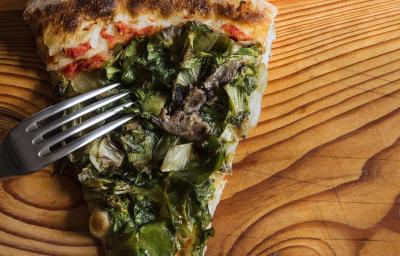 Fotografia em tons de verde em uma bancada de madeira de cor marrom. Ao centro, uma fatia de pizza de escarola com um garfo ao lado. Fotografia em tons de verde em uma bancada de madeira de cor marrom. Ao centro, uma fatia de pizza de escarola com um garfo ao lado.