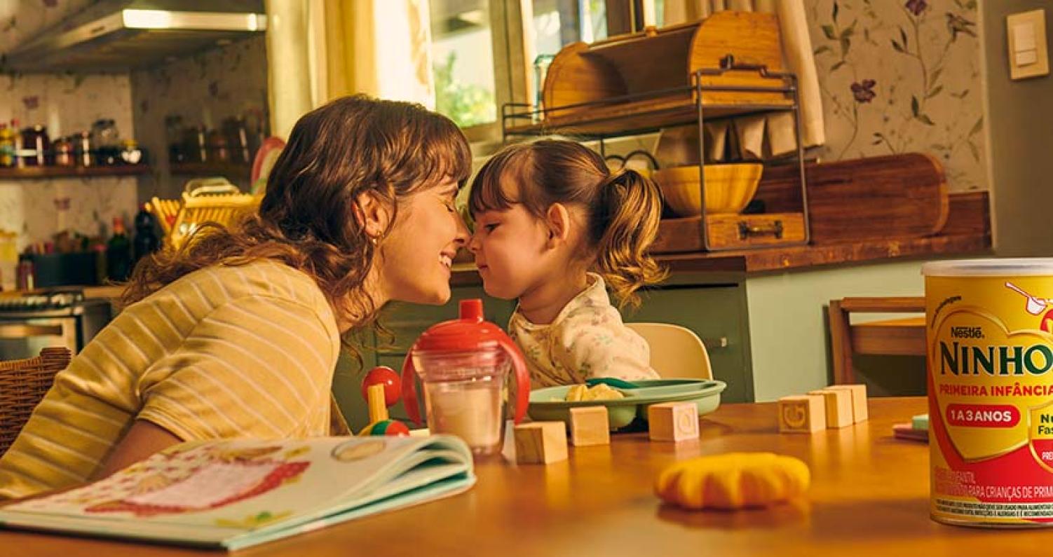 Imagem de uma mãe sorrindo bem perto de sua filha sentadas na mesa de uma cozinha. Em cima da mesa, uma lata de Ninho, alguns