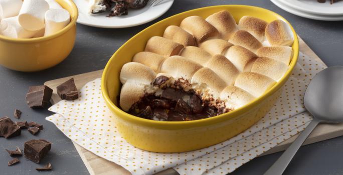 Fotografia em tons de amarelo em uma bancada cinza com um paninho branco de bolinha amarela e um refratário amarelo com o smores de chocolate. Ao fundo, um potinho amarelo com marshmallow e dois pratos com uma fatia do smores em cada um.