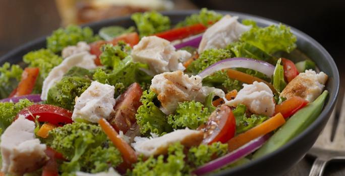 Fotografia em tons de verde, em uma bancada de madeira de cor marrom. Ao centro, um prato redondo preto contendo a salada e ao fundo, um outro prato contendo salada. Ao lado, há um garfo de inox.