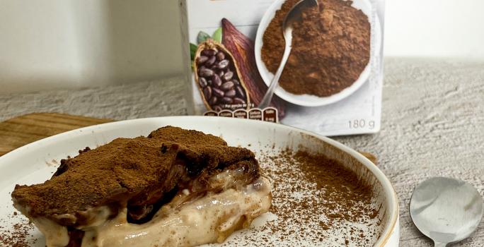 Foto da receita de Bombom de travessa plant-based. Observa-se um pedaço da torta em um prato branco polvilhado de cacau em pó e com com uvas decorando.