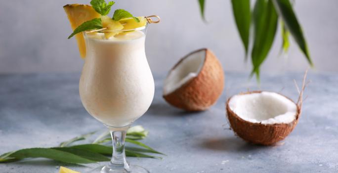 Foto da receita de Piña Colada sem Álcool. Observa-se uma taça grande com a bebida dentro cremosa, uma fatia de abacaxi decorando a borda. Na foto, observa-se um coco seco pela metade.