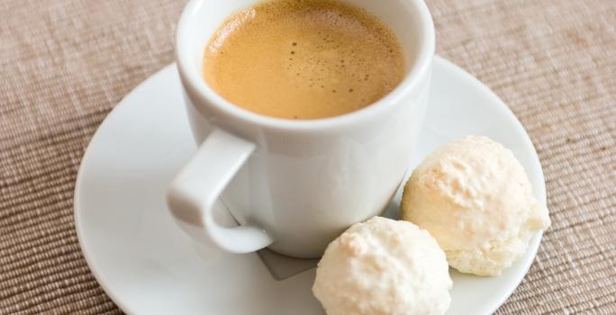 fotografia em tons de branco, cinza e marrom de uma bancada cinza. Contém um pratinho redondo branco com uma xicara de café branca com café dentro, ao lado dois bolinhos redondos.