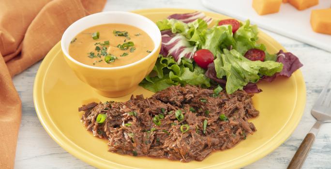 Foto da receita de Purê de Abóbora com Carne Desfiada. Observa-se um prato amarelo com a carne desfiada e a salada e, dentro de um potinho amarelo, o purê de abóbora finalizado com salsinha.