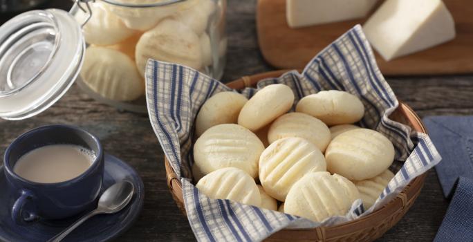 Fotografia em tons de azul em uma bancada de madeira escura, uma cesta de vime com um guardanapo de papel azul listrado com biscoitos de queijo dentro dele. Ao lado, uma xícara de café. Ao fundo, uma tábua de madeira com um pedaço de queijo.