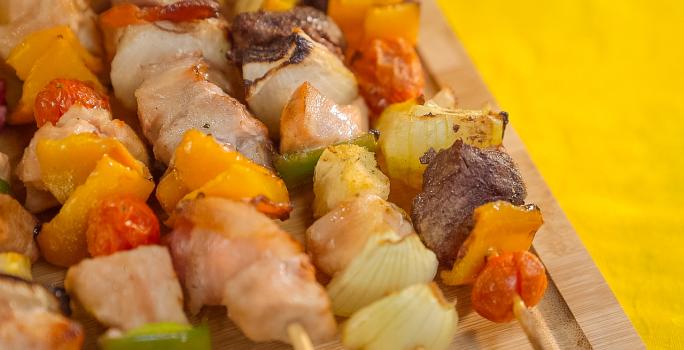 Imagem bem aproximada de uma tábua de madeira com cinco espetinhos sobre ela, alguns com frango, outros com carne e lombo. Eles também têm vegetais em tons verde, amarelo e vermelho.