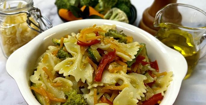 Foto em tons de amarelo da receita de farfalle com legumes servida em uma panela pequena de porcelana vermelha e branca. Ao fundo um vidro de azeite, um triturador de sal, uma porção de legumes e um pote de vidro com farfalle