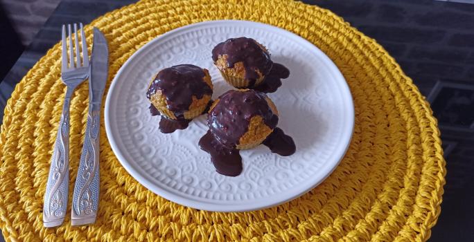 Fotografia em tons de amarelo em uma bancada de pedra, um porta prato amarelo de renda, um prato branco com bolinhos de cenoura com cobertura de chocolate por cima.