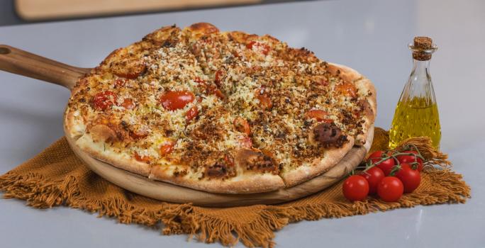 Foto da receita de massa básica de pizza, servida numa tábua de madeira com o sabor de pizza salgada. A tábua está ao lado de um galeteiro de azeite e alguns tomates.