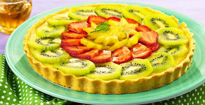 Fotografia em tons de verde e vermelho, com torta de frutas frescas fatiadas em prato verde sobre guardanapo verde com bolinhas brancas, potinho com mel ao fundo, tudo sobre bancada na cor cinza.