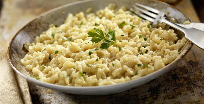 Fotografia de um prato de cor escura com um arroz temperado e salsinha por cima, e ao lado, um garfo apoiado na porta do prato. No fundo da foto, um pão francês cortado na metade, sobre uma toalha de mesa na cor bege.