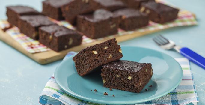 fotografia em tons de azul e marrom de uma bancada vista de frente, contém um pano em tons de branco e azul, um prato azul e redondo contém 2 Brownies e ao fundo um tábua de madeira com diversos Brownies