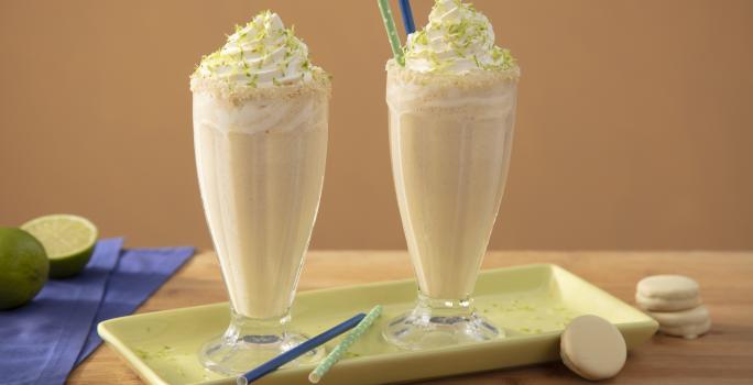 Foto da receita de Milkshake de Bono Torta de Limão Coberto, em dois copos decorados com chantilly, sobre um prato verde, em uma bancada de madeira decora com fatias de limão, biscoitos brancos e um tecido em tom azul