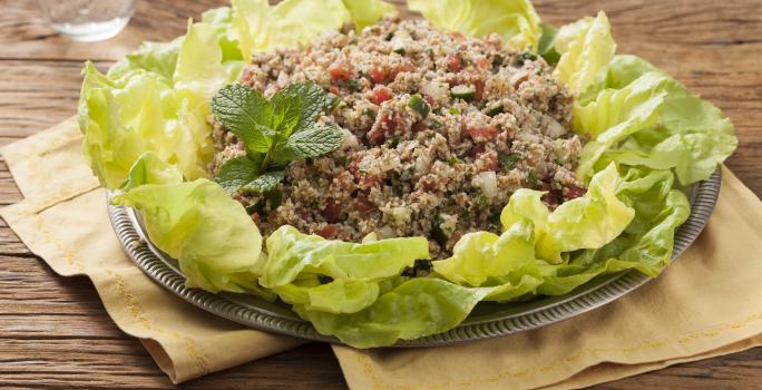 Em uma mesa de madeira contém panos em tons pardo abaixo de um recipiente que comporta a salada para quibe.