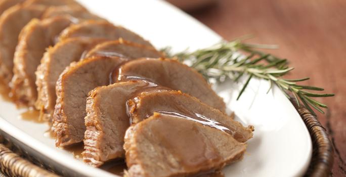 foto em tons marrom e branco. Uma mesa de madeira contém um cesto com um prato que comporta pedaços de carne fatiados.