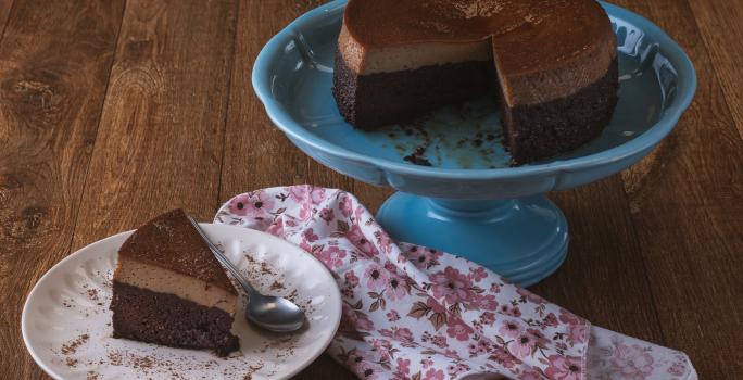 Foto da receita de bolo chocopudim da maria carpio, servido sobre um prato grande azul, com uma fatia cortada sobre outro prato branco, tudo em uma bancada de madeira