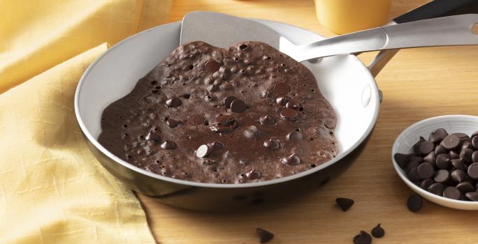 Fotografia em tons de amarelo em uma bancada amarela e um pano amarelo na lateral. Uma frigideira ao centro com o brownie de chocolate dentro dela. Ao lado, um potinho com gotas de chocolate.