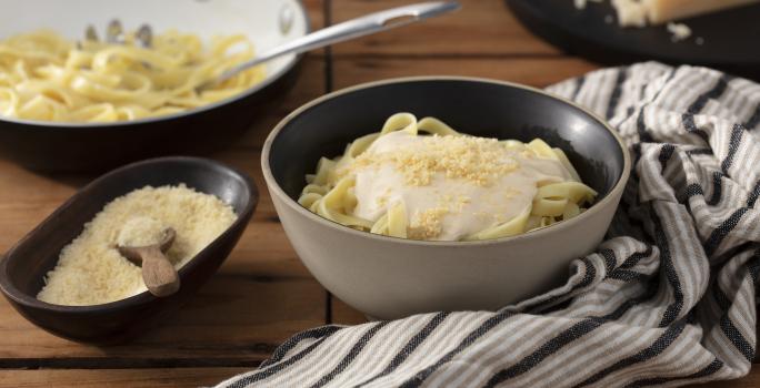 Fotografia em tons de marrom em uma bancada de madeira escura, um pano branco com listras marrom, um recipiente redondo fundo bege com o fettuccine com molho alfredo e queijo ralado por cima dentro dele. Ao lado, um potinho com queijo ralado.