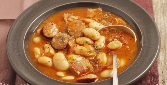 Fotografia de um prato fundo na cor preta com uma sopa de feijão branco e linguiça. Ao lado, três fatias de pão e do outro lado, um tempero em pó vermelho dentro de um pote pequeno branco de cerâmica, que está em cima de um pano em tons de vinho.