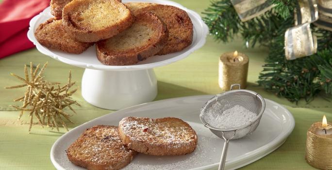 Foto da receita de rabanada sem glúten servida sobre uma base de porcelana branca com duas fatias separadas polvilhadas com açúcar de confeiteiro. Ao fundo há um paninho vermelho e decoração natalina