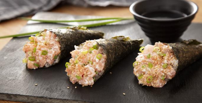 Foto da receita de Temaki de Salmão. Observa-se 3 temakis em cima de uma tábua de preta e, atrás, um potinho com molho shoyu e um ramo de cebolinha.