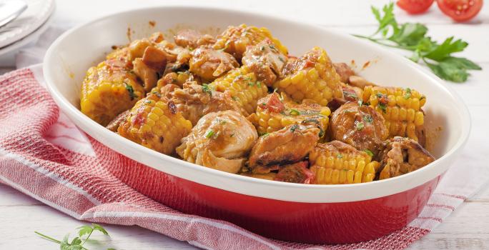 Fotografia em tons de amarelo, vermelho e branco de uma bancada de madeira branca com paninho vermelho, sobre ele um recipiente oval vermelho com bordas brancas com pedaços de frango e milho. Ao fundo duas fatias de tomate e um prato redondo branco.