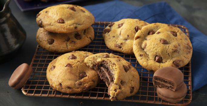 Fotografia em tons de preto e azul de uma bancada preta com um grade dourada, sobre ela cookies. Ao fundo um paninho azul e biscoitos negresco cobertos de chocolate espalhados.