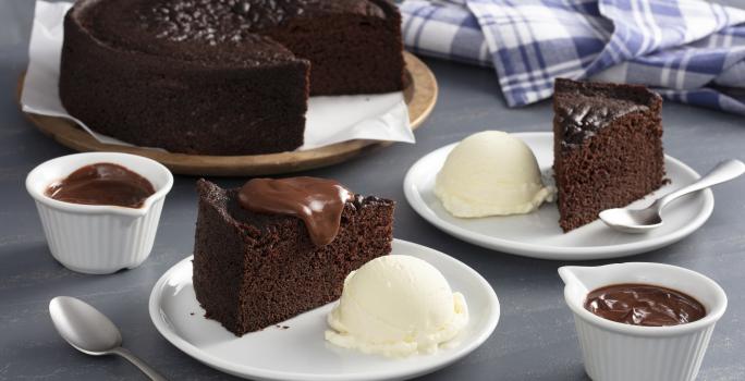 Fotografia em tons de marrom e azul de uma bancada cinza, ao centro dois pratos brancos com fatias de bolo de chocolate e ao lado uma bola de sorvete de creme. Ao fundo o bolo inteiro sem duas fatias.