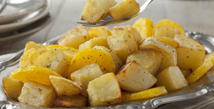 Fotografia em tons de amarelo em uma bancada de madeira com um recipiente de prata com várias mandioquinhas salteadas.