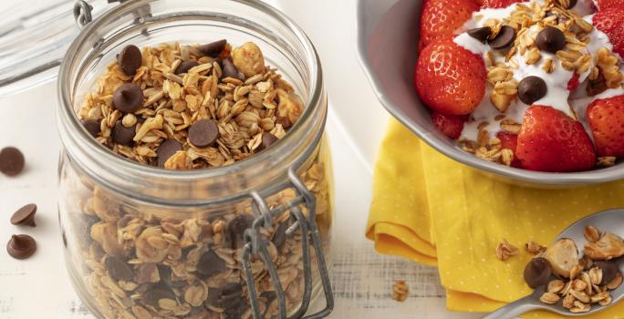 Fotografia em tons de branco de uma bancada branca com um pote de vidro com tampa  e granola. Ao lado um prato branco com paninho amarelo e uma salada de frutas com granola.