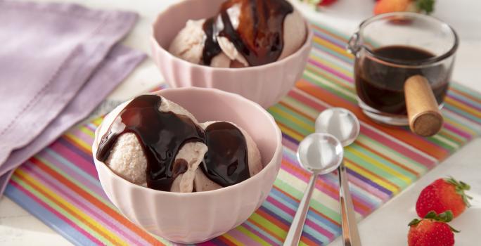 Foto da receita de Sorvete de Morango Caseiro com Calda de Leite Moça e Nescau. Observa-se dois recipientes com duas bolas de sorvete e a calda de chocolate por cima. Morangos decoram a foto.