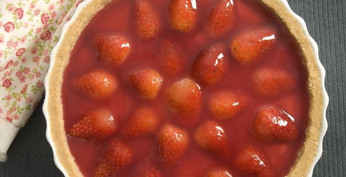 Fotografia em tons de vermelho com em uma bancada de madeira clara com um pano vermelho, um prato branco raso grande com a torta de morango.