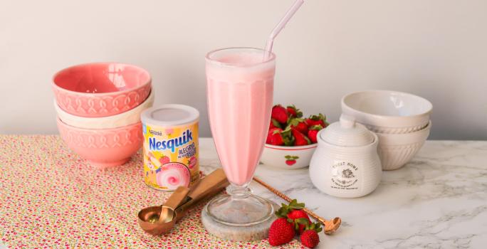 Fotografia de um milkshake rosa servido em uma copo alto de vidro. Ao fundo um cenário com morangos e bowls com tons rosas.