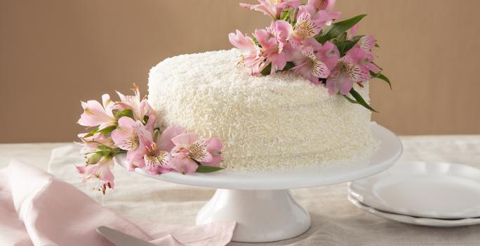 Foto da receita de Bolo de Gala. Observa-se um bolo grandioso com flores rosas sobre uma boleira de cerâmica branca. Atrás, um fundo nude e pratinhos de sobremesa decoram a foto, assim como um guardanapo rosa.