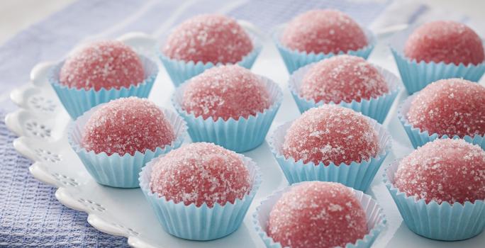 Fotografia em tons de azul e rosa de um paninho azul, sobre ele um prato redondo com decoração de florezinhas, sobre ele 12 brigadeiros rosa, com açúcar polvilhado, em forminhas azuis claras para doce.