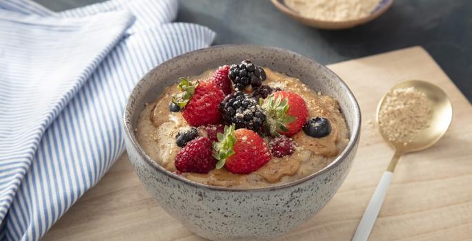 Foto em tons de bege da receita de mingau nutritivo servida em um bowl de cerâmica sobre uma tábua de madeira clara com uma colher prateada ao lado. Ao lado ainda há um paninho azul listrado e um recipiente com aveia em flocos