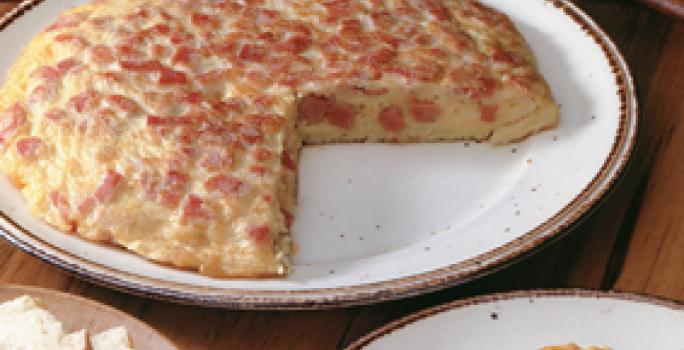 Fotografia em tons de vermelho em uma mesa de madeira com um prato branco raso grande com a omelete cortada ao meio. Ao lado, um prato com a fatia da omelete e outro prato com fatias de pão.