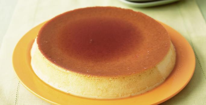 Fotografia de um pudim com calda de caramelo por cima, servido em um prato amarelo sobre uma mesa de madeira
