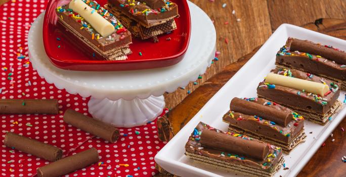 Foto de uma bancada de madeira, sobre ela há um pano vermelho com bolinhas brancas, um prato retangular com a receita pronta, alguns chocolates Baton soltos e mais um prato vermelho elevado. Há alguns granulados coloridos decorando a cena.