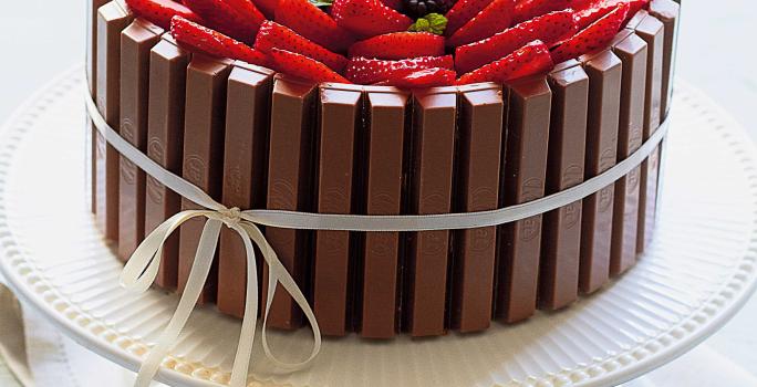 Fotografia em tons de branco de uma bancada branca com um paninho branco, um suporte para bolo branco e um bolo de Kitkat com frutas vermelhas. Ao lado um recipiente redondo branco com frutas vermelhas.
