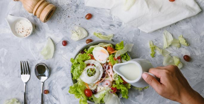 Fotografia vista de cima de uma mão segurando uma jarra pequena na cor branca, que está caindo um molho branco em uma salada de folhas, cebola, ovo cozido, maçã, tomates cereja, cenoura e azeitona preta. O refratário está sobre uma mesa de mármore branca.