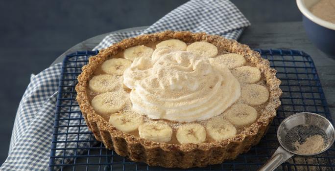 Foto da receita de banoffee de farinha lactea aveia servida em uma porção grande sobre uma grade preta com uma colher de servir prateada ao lado