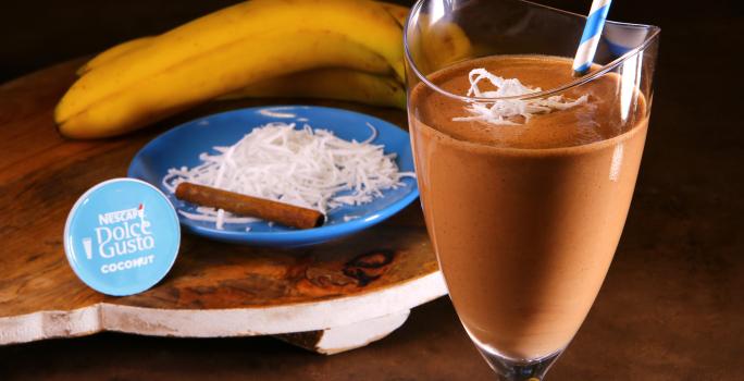 Fotografia em tons de azul em uma bancada de madeira, uma tábua de madeira com um copo de vidro alto e a bebida de chocolate com coco e banana dentro dele. Ao fundo, um prato azul com raspas de coco, uma canela em pau e uma banana.