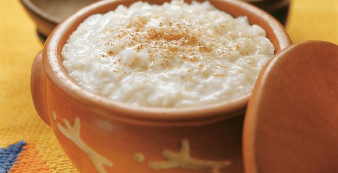 Fotografia em tons de amarelo em uma mesa de madeira com uma toalha amarela desenhada, um recipiente de barro com o arroz doce dentro dele. Ao fundo, um potinho com paus de canela.