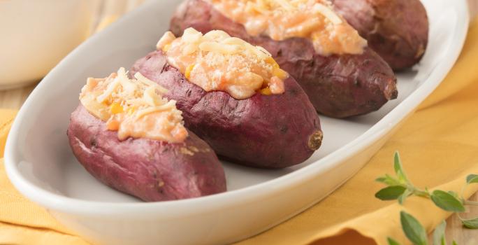 Fotografia em tons de amarelo em uma bancada de madeira com um pano amarelo, um recipiente oval branco com as batatas-doces recheadas com frango cremoso.