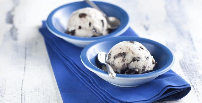 Fotografia em tons de azul em uma bancada de madeira clara, um pano azul, dois potinhos azuis redondos com o sorvete feito com Biscoito Negresco.
