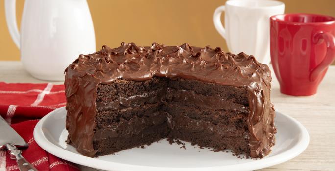 Fotografia em tons de vermelho em uma bancada de madeira com um pano vermelho listrado em branco, com um prato branco no centro com o bolo brownie com brigadeiro de cacau de cobertura. Ao fundo, uma xícara branca, uma xícara vermelha e um bule branco.