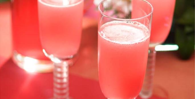 Fotografia em tons de rosa em uma bancada de madeira, um pano vermelho, taças de vidro de champagne com as bebidas feitas com Picolé de Limão com espumante.